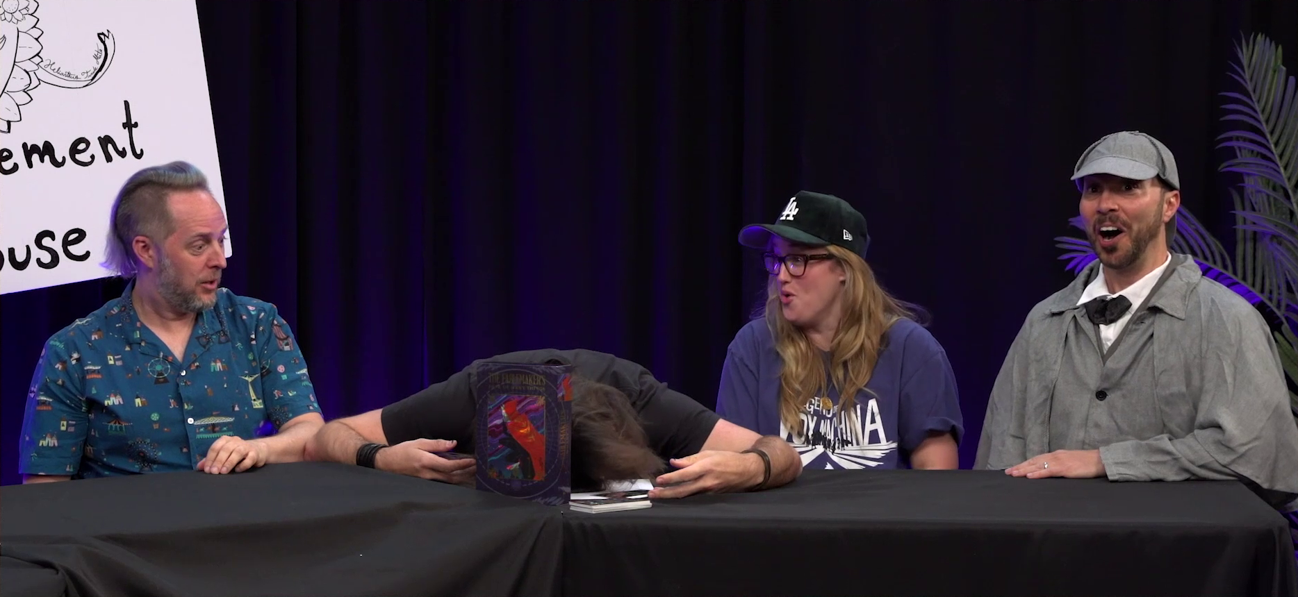 Taliesin, Matt, Ashley, and Sam at the Announcement Playhouse table. Matt is face down on the table, apparently dead. Taliesin and Ashley look at Matt with surprise and worry. Sam, dressed as Sherlock Holmes in a deerstalker hat and Inverness cape, looks directly into the camera, his mouth open in astonishment. On the table in front of Matt is a box with a caped individual on the front labeled “THE FABLEMAKERS”. A deck of cards sits next to the box.