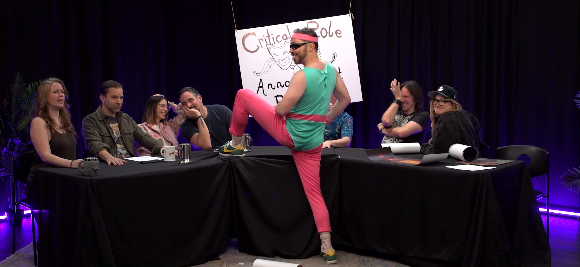 The Critical Role Announcement Playhouse table. Sam wears a neon pink and green 80’s-style exercise suit and stands with his left leg up on the table, crotch pointing towards the rest of the cast. Marisha looks confused, Liam is holding back a smirk, Laura and Travis look at each other laughing, Matt grimaces and covers one eye, and Ashley purses her lips in mock discomfort. Sam wears a pink headband and sunglasses. The Announcement Playhouse sign bears a hand-drawn image of a laughing cat drama mask.