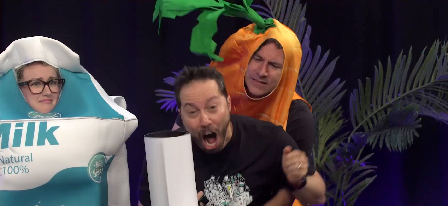 The Critical Role Announcement Playhouse table. Matt, dressed as a carrot, performs the Heimlich maneuver on Sam, who gasps violently, appearing to expell a large rolled up piece of poster paper. Ashley, dressed as a carton of milk, watches, looking disturbed.