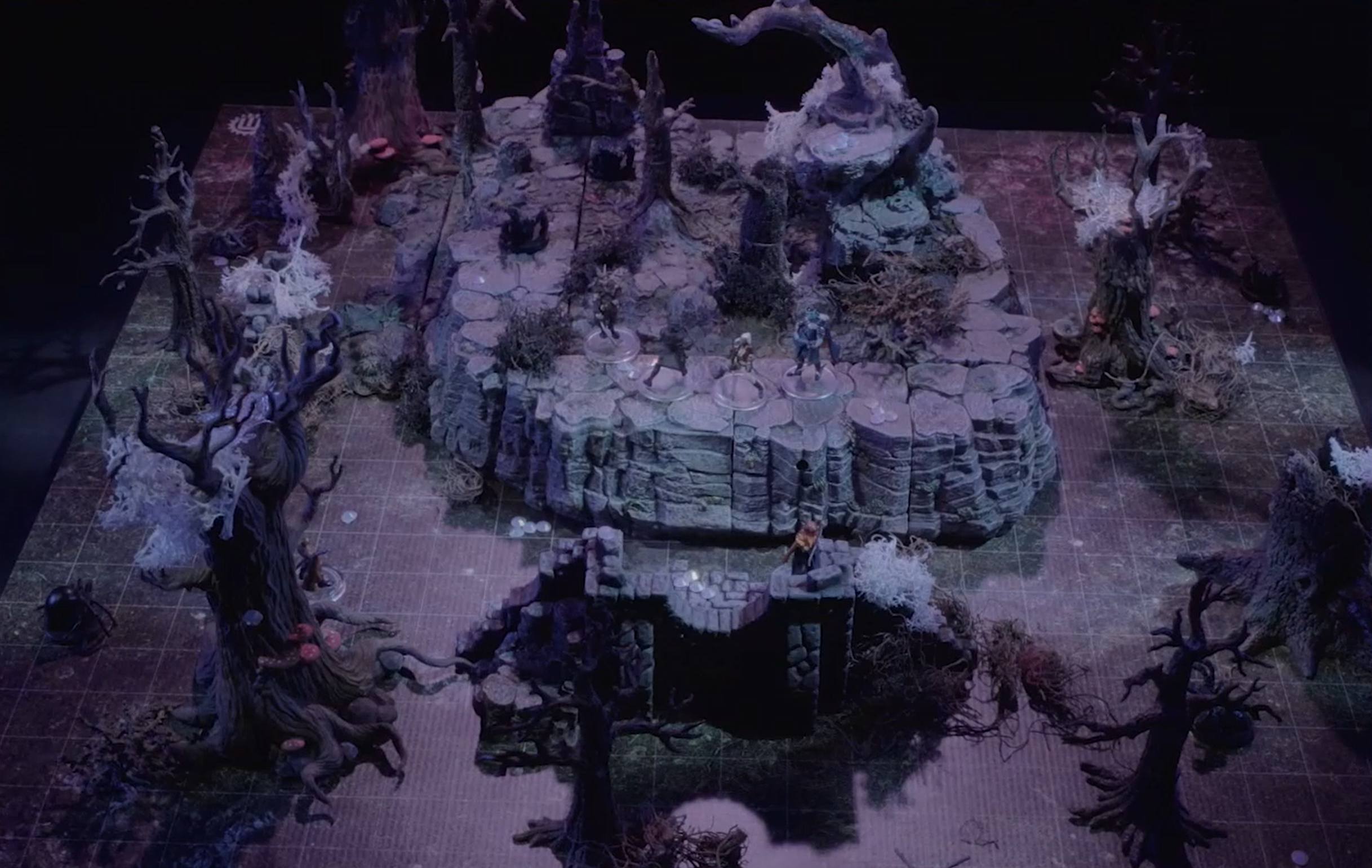 A battlemap of a rocky, forested area. Leafless trees covered in white web-like growths and mushrooms surround the ruins of a wall and a large rocky mound. A number of groups of crystals in the vague shape of penises are scattered about. Opal and a shadowy Ted face off against Morrighan and Dorian on the mound while Dariax and Fy’ra stand at ground level.