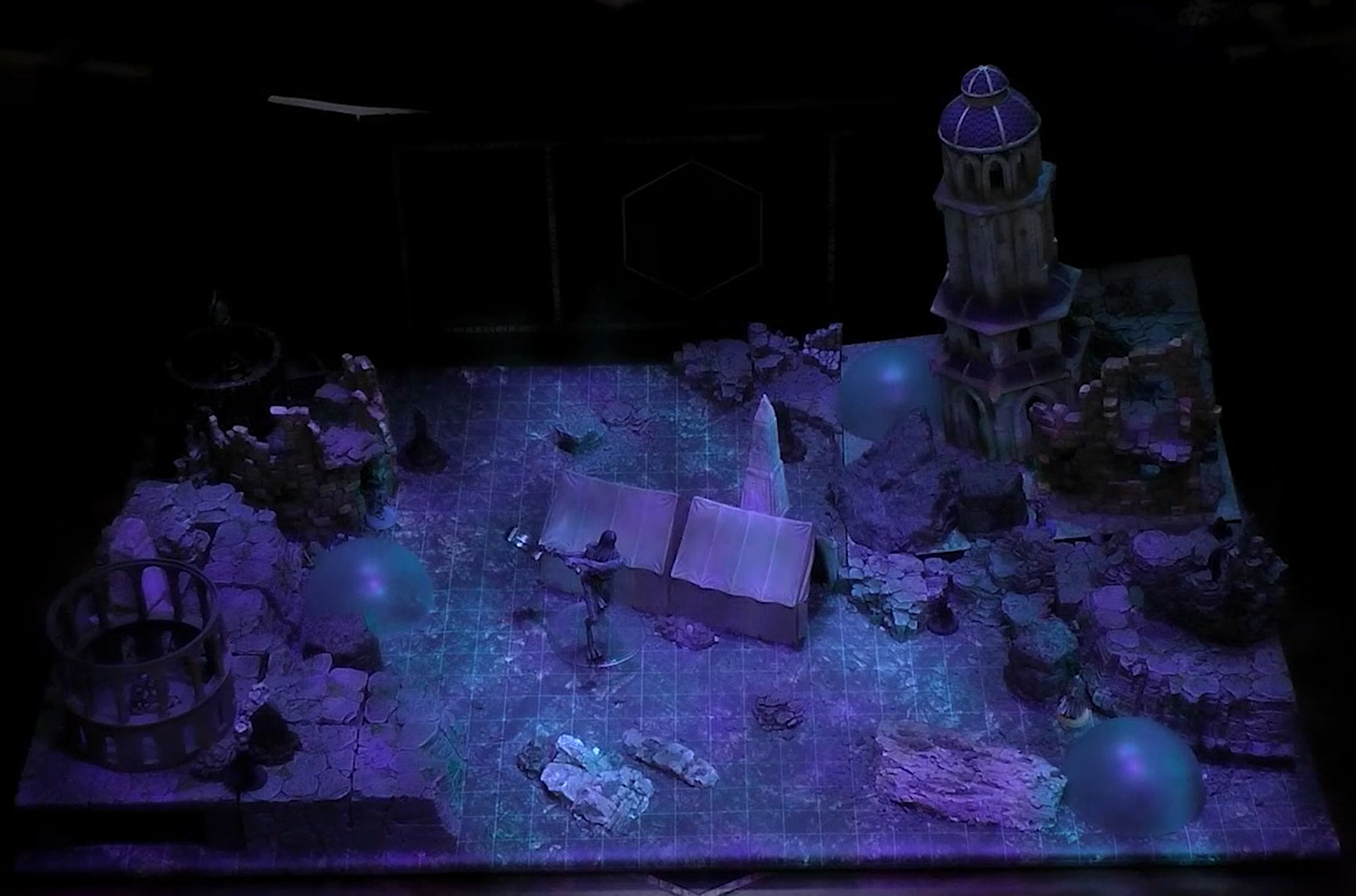 A battlemap of a dark underground cavern filled with the ruins of a civilization. In the center next to a pale white obelisk and two rectangular tents is a tall, thin, gray creature with folds of skin hanging from its face wielding a hammer. Further out a large tower stands among crumbling walls and three blue translucent domes.