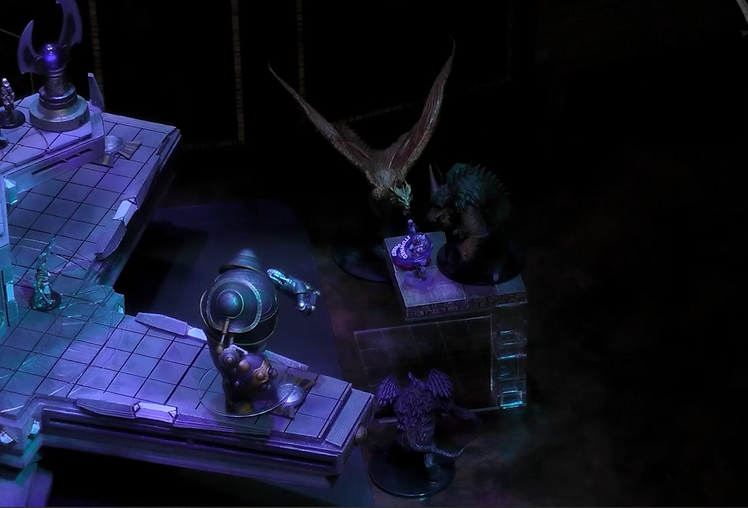 A close-up of a battlemap, focused on the end of a square walkway next to a rotating four-tipped platform. The Emissary stands surrounded by demons and a flying dragon, with a bulbous robotic Irrevocable construct on the tip of the rotating platform next to him. His miniature has two rings which say “STUNNED” around it.