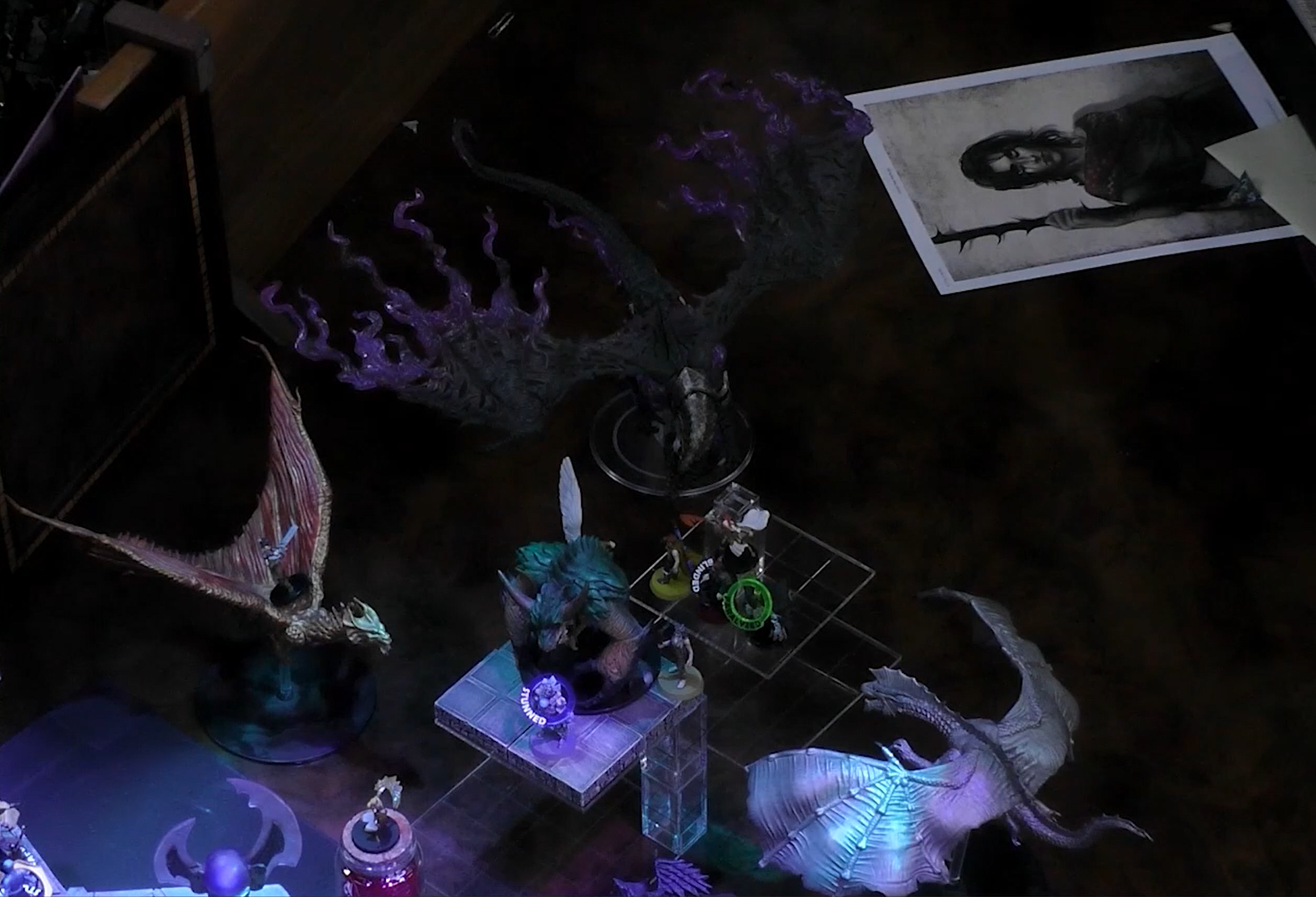 A close-up of a battlemap, focused on the end of a square walkway where the party faces off against a number of enemies, including three dragons: a brass dragon with red streaks in its wings, a silver dragon with a bony crest, and a shadowy-black dragon whose wings taper off into purple flame. Tishar rides the brass dragon. A print-out of Asha’s character art is off to the side.