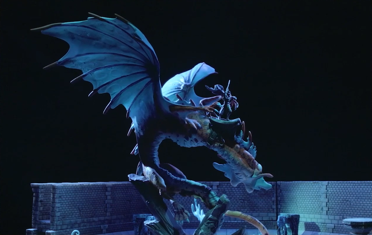 A miniature of Gloamglut, a massive snake-like dragon, stands in the middle of a stone chamber. Upon its back is Sorrowlord Zathuda in black armor holding a sword aloft. Gloamglut’s wings are outstretched and its fish-like head looks off to the side.