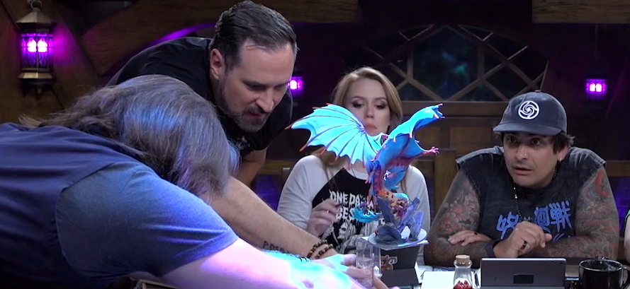 Travis helps Matt position a miniature of Gloamglut, a massive snake-like dragon with outstretched wings. Marisha looks in interest while Robbie stares transfixed, his mouth partially open.