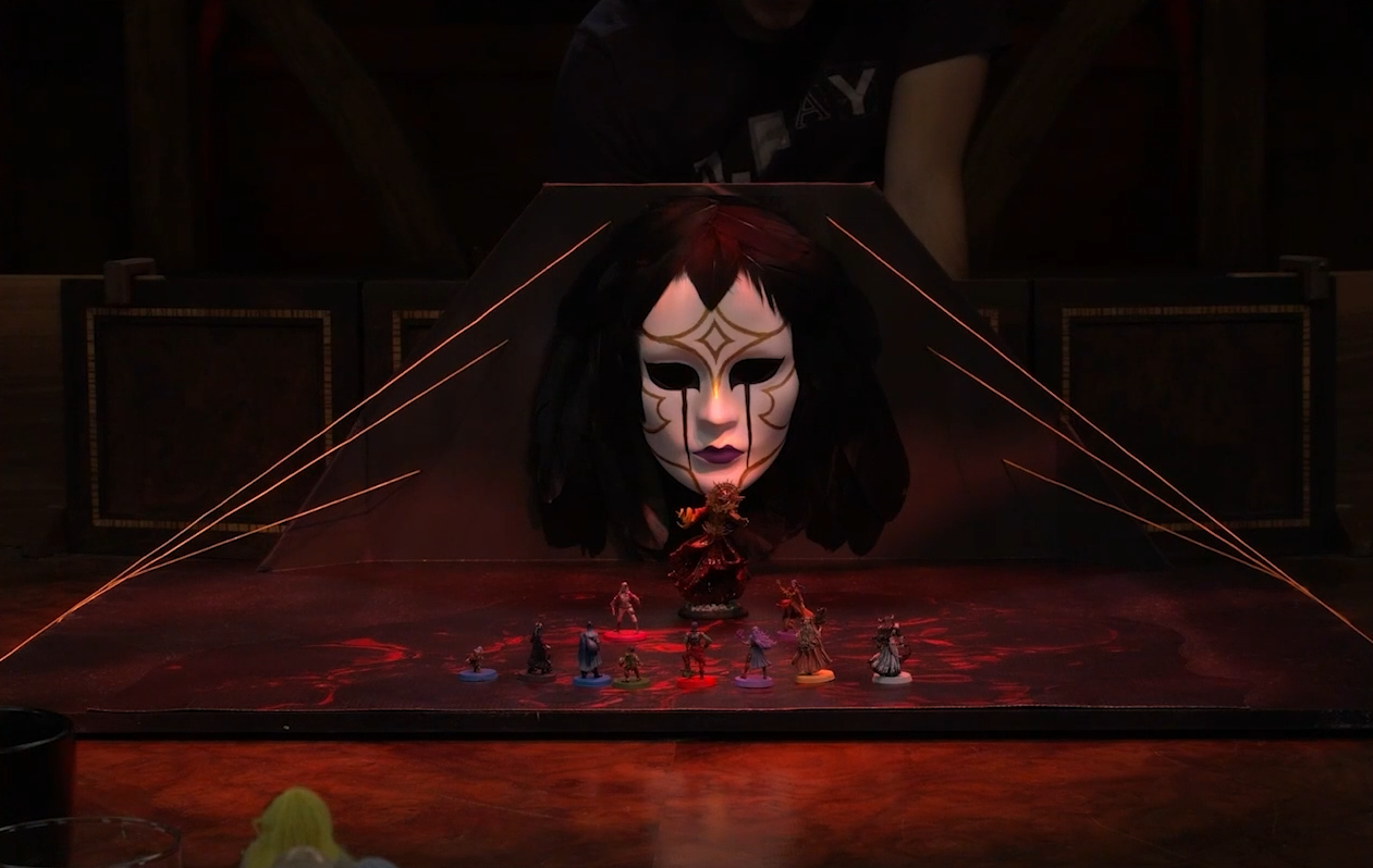 A battlemap depicting a floor of glowing red blood looked over by a patterned porcelain mask with hair of black feathers. Chetney, Laudna, Dorian, Orym, Ashton, Imogen, Fearne, and Braius stand in a line facing off against Opal, Liliana, and a giant Vespin Chloras between them. Six golden strands run from behind the mask to the edges of the floor. Matt stands in the background.