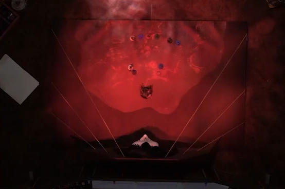 A top-down view of a battlemap depicting a floor of glowing red blood looked over by a patterned porcelain mask with hair of black feathers. Chetney, Laudna, Dorian, Orym, Ashton, Imogen, Fearne, and Braius stand in a line facing off against Opal, Liliana, and a giant Vespin Chloras between them. Six golden strands run from behind the mask to the edges of the floor.