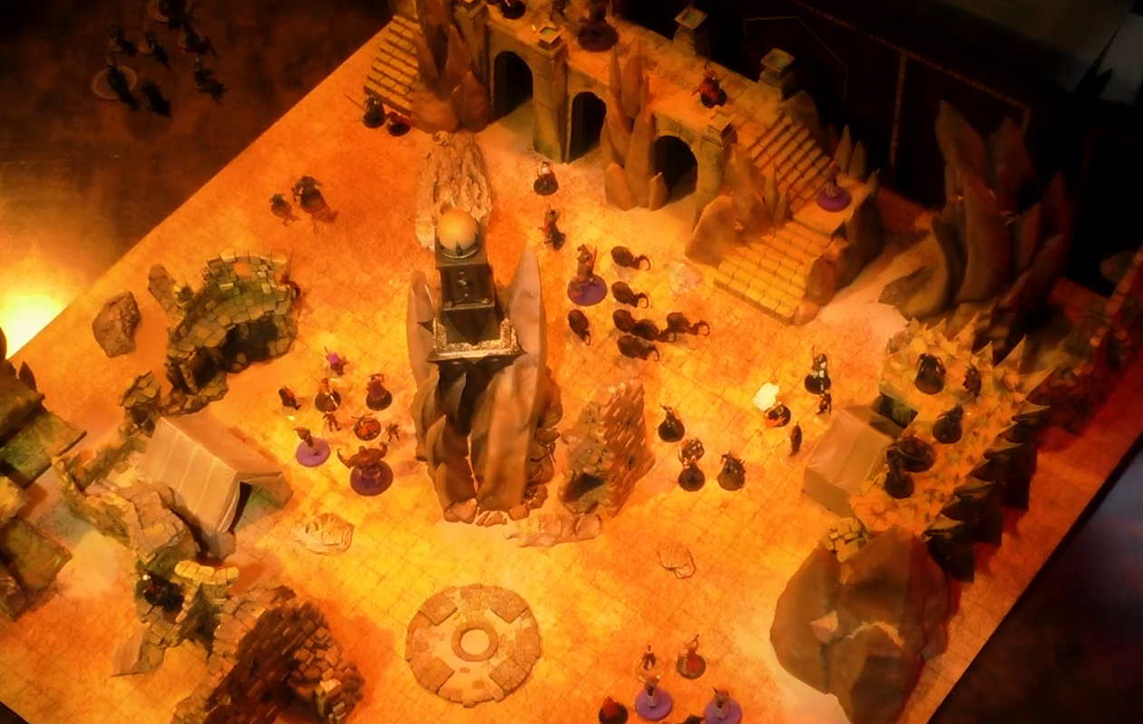 A battlemap of a large outdoor space filled with stone walls and parts of buildings. In the center is a tower with a pale white globe at the top. A number of combatants fill the area on the ground and atop the walls.