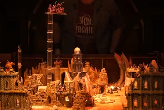 A battlemap of a large outdoor space filled with stone walls and parts of buildings. In the center is a tower with a pale white globe at the top. A number of combatants fill the area on the ground and atop the walls. A set of tokens is floating high above the battlefield. Matt, wearing a Vox Machina T-shirt, is visible in the background.