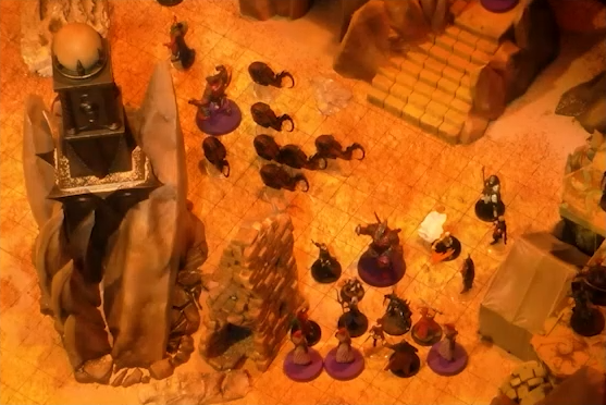 A battlemap of an outdoor space filled with stone walls and parts of buildings. A tower with a pale white globe at the top is off to the side. A number of combatants face off on the ground, including a collection of slithers around a large Reiloran.