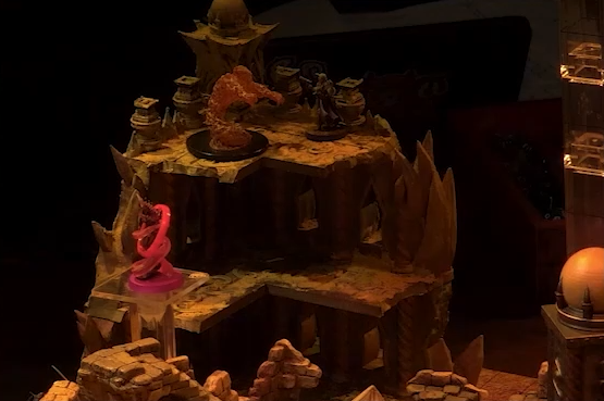 A battlemap focusing on a tiered section of wall. A fire elemental stands at the top level, facing off against a combatant. A Reiloran surrounding by swirling red energy is on the level below. A tower with a pale white globe at the top is just at the edge of the frame.