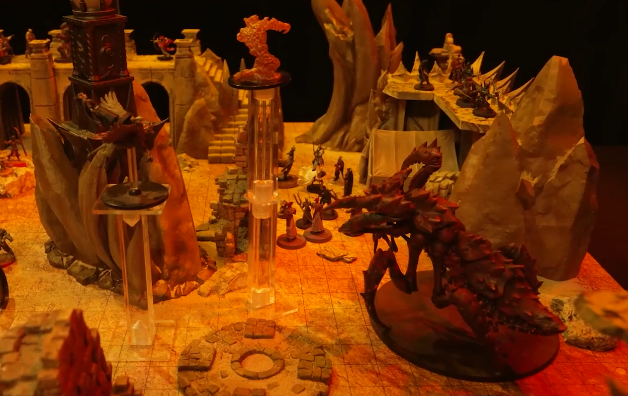 A battlemap of a large outdoor space filled with stone walls and parts of buildings. A number of combatants fill the area on the ground and atop the walls. A large, red, insectoid vidulch stands off to the side. A fire elemental flies near a table.