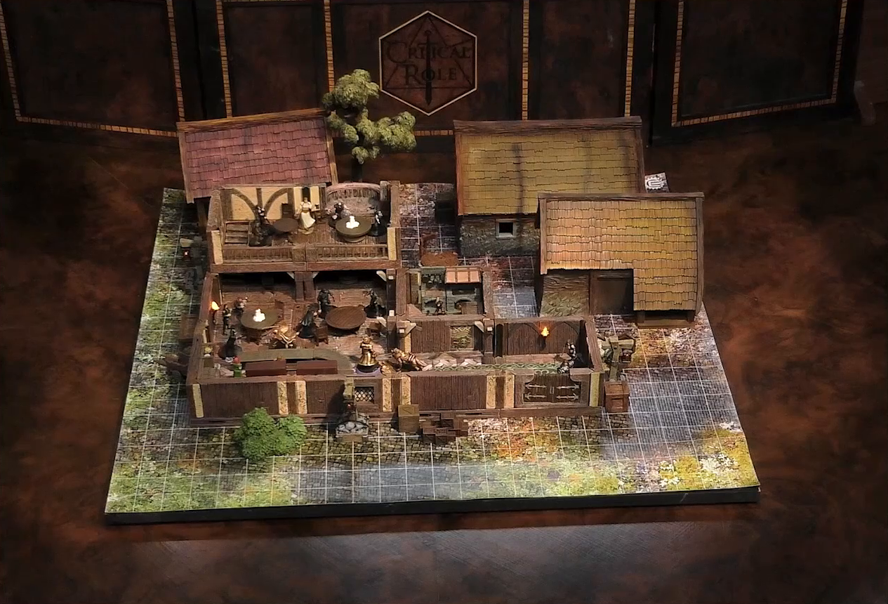 A battlemap of the interior of a wooden tavern with a couple small shed-like buildings next to it. A number of black-cloaked individuals and other rogues stand near round tables, facing off against Thimble, Occtis, Kattigan, and Wulfric. An upper level of the tavern has more tables, rogues, and an individual in a white dress who appears to be a server. A small side room houses brick stoves.
