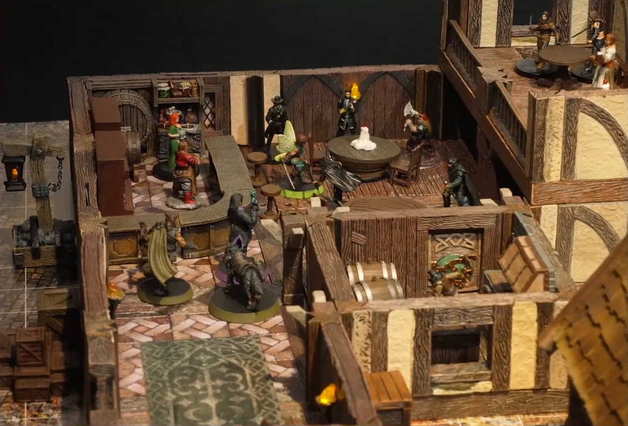 A battlemap of the interior of a wooden tavern. A number of black-cloaked individuals and other rogues stand near round tables, facing off against Thimble, Occtis, Kattigan, and Wulfric. Two bartenders, one thin and in a green dress, one stout and burly, stand behind a countertop. A small side room houses brick stoves, barrels, and a long-armed, beast-like individual. An upper level of the tavern has more tables, rogues, and an individual in a white dress who appears to be a server. A lit lantern hangs from a post outside the tavern.