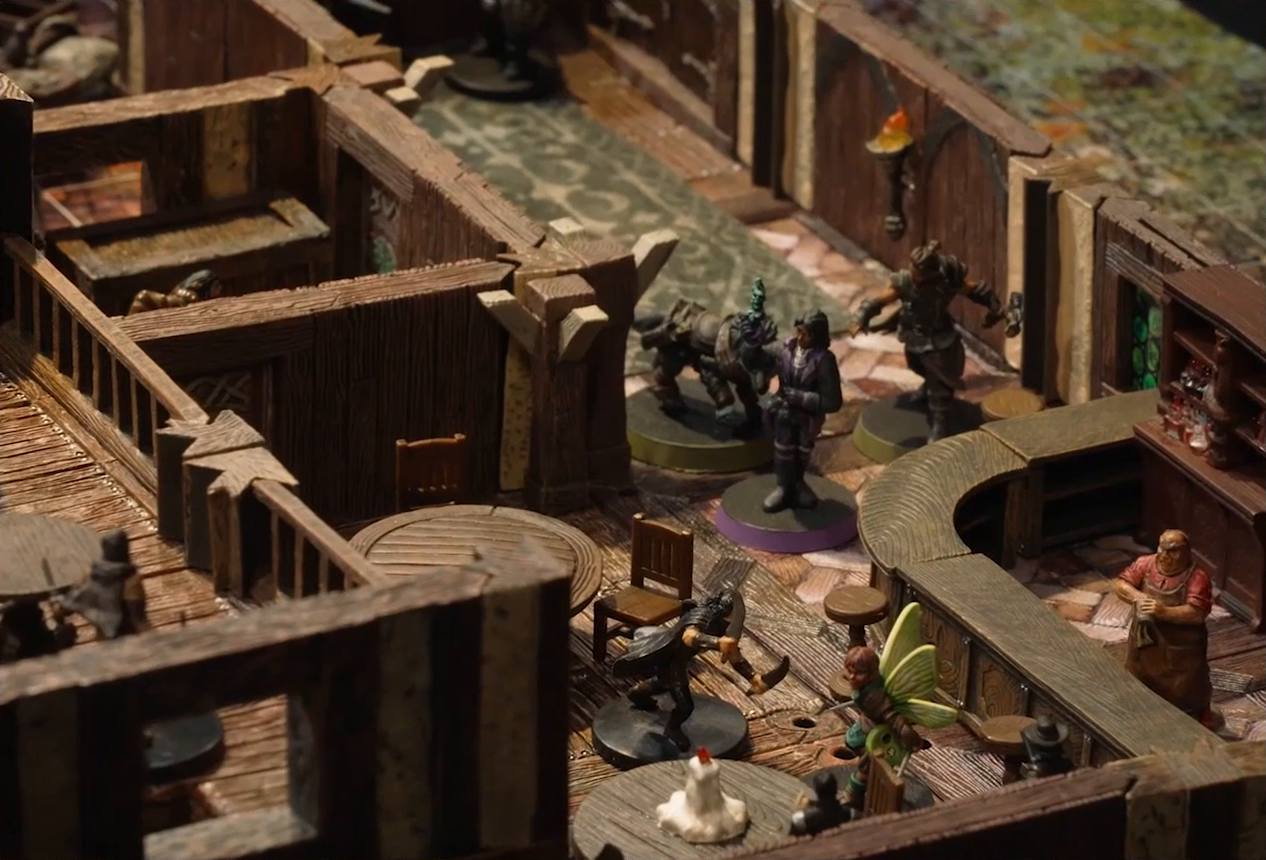 A battlemap of the interior of a wooden tavern. Thimble faces off against a black-cloaked individual wielding daggers while Occtis, Kattigan, and Wulfric approach from down the hall. Some round tables sit near the fight, one with a melting white candle in the center. A bartender stands behind a counter, cleaning a glass. Another rogue can be seen on an upper level, one in a small side room, and one at the end of a hallway near the tavern entrance.