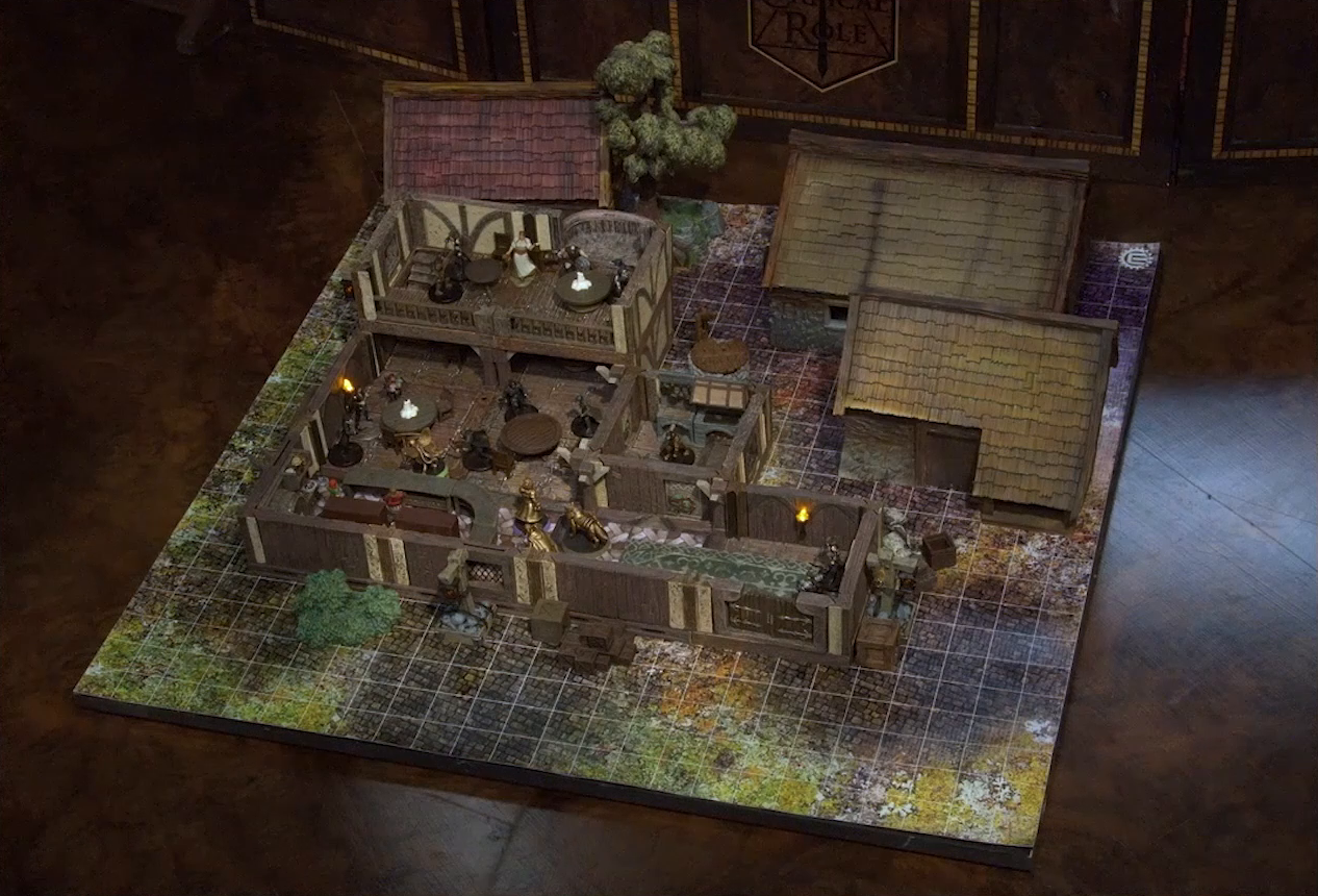 A battlemap of the interior of a wooden tavern. A number of black-cloaked individuals and other rogues stand near round tables, facing off against Thimble, Occtis, Kattigan, and Wulfric. Two bartenders, one thin and in a green dress, one stout and burly, stand behind a countertop. A small side room houses brick stoves, barrels, and a long-armed, beast-like individual. An upper level of the tavern has more tables, rogues, and an individual in a white dress who appears to be a server. A covered well sits outside the tavern.