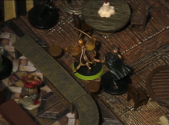 A battlemap of the interior of a wooden tavern focused on Thimble facing off against a black-cloaked individual wielding daggers. A bartender stands behind a counter, cleaning a glass. Another rogue is just out of frame near a round table with a melting white candle in the center.
