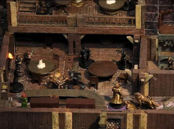 A battlemap of the interior of a wooden tavern. A number of black-cloaked individuals and other rogues stand near round tables, facing off against Thimble, Occtis, Kattigan, and Wulfric. Two bartenders, one thin and in a green dress, one stout and burly, stand behind a countertop. A small side room houses brick stoves, barrels, and a long-armed, beast-like individual. An upper level of the tavern has more tables and rogues.
