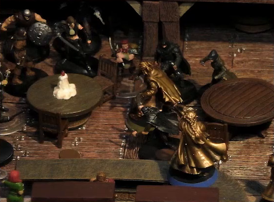A battlemap of the interior of a wooden tavern focused on Kattigan and Vaelus facing off against a number of black-robed individuals and other rogues. They fight amongst round tables, one of which has a melting white candle in the center. Two bartenders watch from the other side of a counter, on which Vaelus stands.