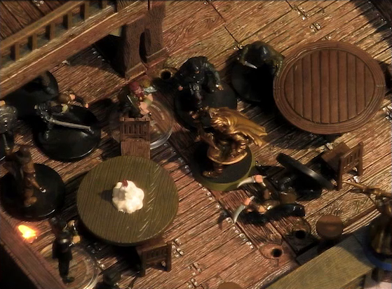 A battlemap of the interior of a wooden tavern focused on Kattigan facing off against a number of black-robed individuals and other rogues. One rogue next to Kattigan lays on the ground. They fight amongst round tables, one of which has a melting white candle in the center. Vaelus stands on a counter at the edge of the image. The railing of an upper level is visible in the background.
