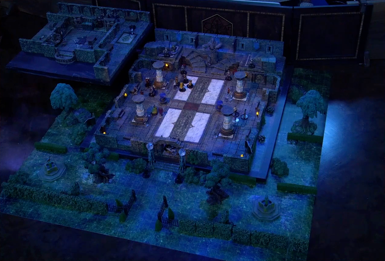 A battlemap of a stone palace split into two levels, surrounded by a courtyard. Downstairs is a marble floor, marble pillars, and twin staircases curving up on either side of a large robed statue holding a lantern and open book. A number of people are scattered throughout the downstairs space. Upstairs is a hallway leading past a room with a bearskin rug, wooden table, and balcony. A number of robed individuals including Ethrand Tachonis gather outside the room, down the hall from a fallen guard. Two black-clothed individuals stand outside the entrance to the palace next to two pillars topped with large spherical crystals. The rest of the courtyard contains trees, two fountains, and an open gate leading in.