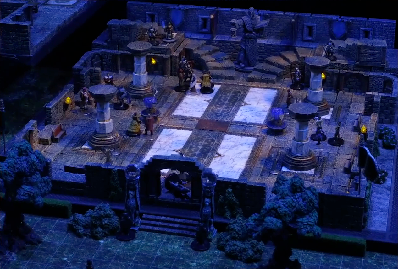 A battlemap of a stone palace split into two levels. Downstairs is a marble floor, marble pillars, and twin staircases curving up on either side of a large robed statue holding a lantern and open book. A number of people are scattered throughout the downstairs space. The edge of the upstairs space is just visible at the left side of the image. Two black-clothed individuals stand outside the entrance to the palace next to two pillars topped with large spherical crystals, and someone lays on the ground in the palace doorway.