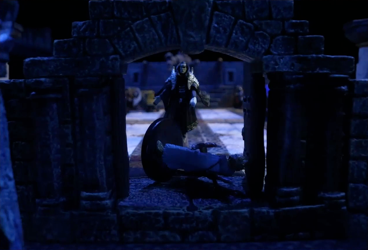 A battlemap of a stone palace focused on the entrance. A stony-faced Primus Tachonis, wearing long black robes and silver shoulder armor, stands in the entryway over a fallen guard. Behind him inside the palace is a marble floor and a number of scattered individuals.