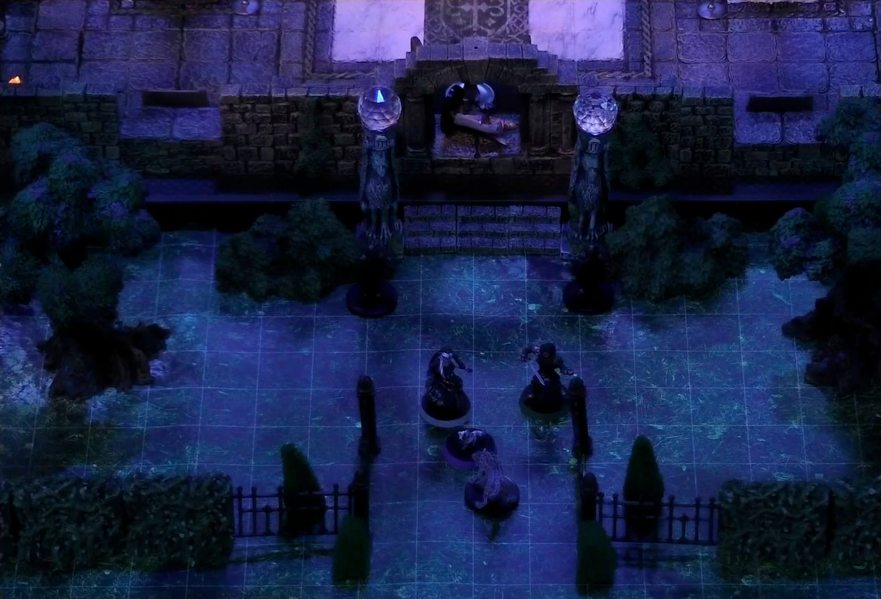 A battlemap of a stone palace surrounded by a courtyard. Vaelus, Julien, Pincushion, and Thaisha in wolf form stand outside the palace entrance, just inside the open gate. Two black-clothed individuals stand outside the entrance to the palace next to two pillars topped with large spherical crystals. Primus Tachonis stands just inside the entrance over a fallen guard.