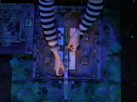 A top-down view of a battlemap of a stone palace split into two levels, surrounded by a courtyard. Brennan’s arms, wearing black-and-white-striped mesh, hold a long transparent measurement tool from the center of the palace out the front door reaching Vaelus, Julien, Pincushion, and Thaisha in wolf form standing outside. Downstairs is a marble floor, marble pillars, and twin staircases curving up on either side of a large robed statue. A number of people are scattered throughout the downstairs space. Upstairs is a hallway leading past a room with a bearskin rug, balcony, and wooden table on which Occtis lays, surrounded by robed individuals.