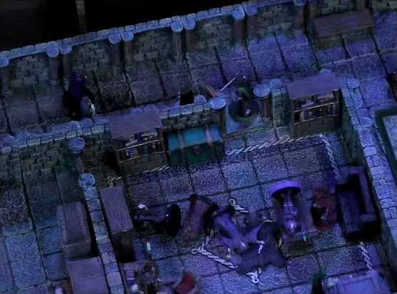 A battlemap of a stone palace upper floor. Occtis lays on a wooden table in a small room with a bearskin rug. Thaisha stands over him and Julien guards the doorway. A number of robed individuals lay on the ground in the room. Two more individuals stand in the hallway.
