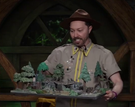 Sam carefully carrying a battlemap of a forest clearing with a bridge running down the center. Sam wears a tan scout uniform and brown brimmed campaign hat.