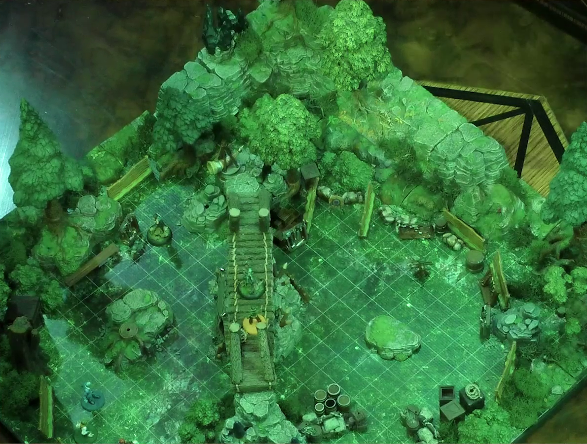 A battlemap of a forest clearing cut down the center with a log bridge spanning two stone ledges. Rocky outcroppings border two sides of the map. A number of boxes, crates, and wooden barriers are strewn throughout. Beau stands in the center of the bridge facing off against Kai. Yasha, Jessep, Padmund, and Sky are on the ground to the left of and below the bridge.