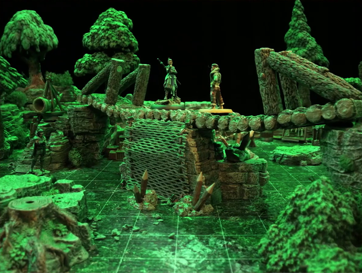 A battlemap of a forest clearing cut down the center with a log bridge spanning two stone ledges. Beau stands in the center of the bridge facing off against Kai. Yasha stands on the ground to the side of the bridge. A net of climbable-looking ropes leads up from the ground to the bridge’s center, and wooden stakes sprout from its base.