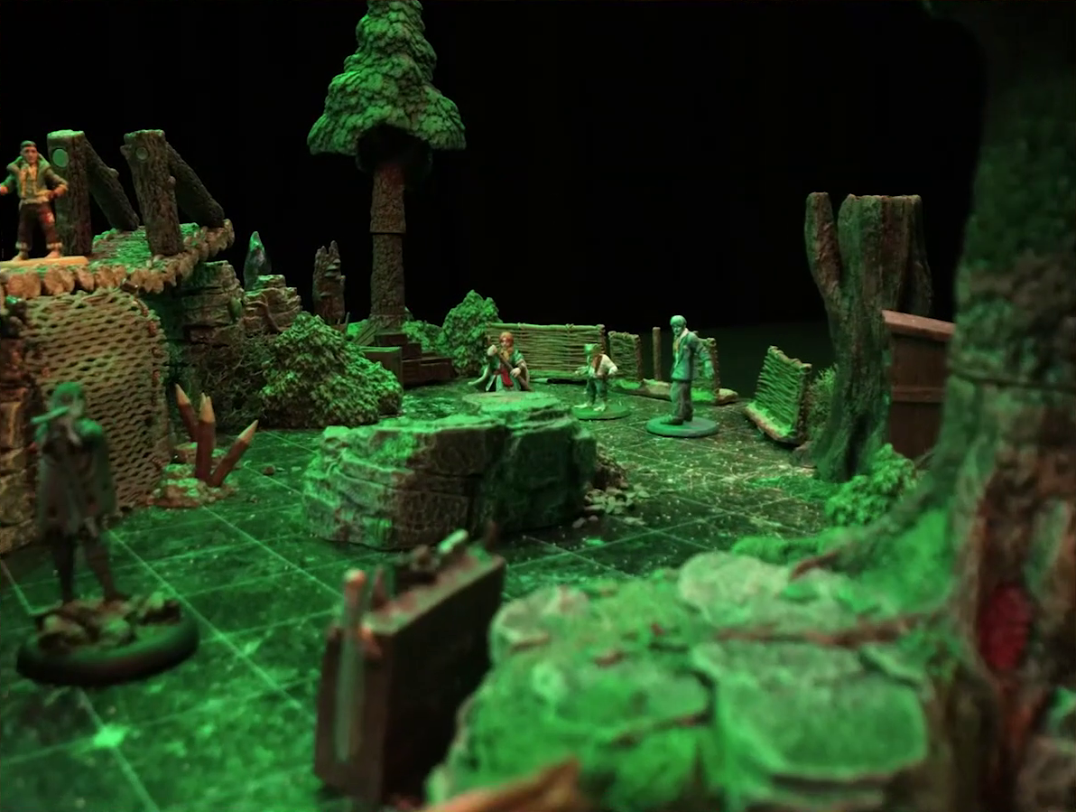 A battlemap of a forest clearing. Sky, Padmund, and Jessep stand in a line behind a rocky outcropping and in front of a wooden fence, while Yasha is visible but blurry in the foreground. To the left of the frame is a log bridge raised above the forest floor on which Kai is standing. The bridge begins at a stone ledge. A net of climbable-looking ropes leads up from the ground to the bridge’s center, and wooden stakes sprout from its base.