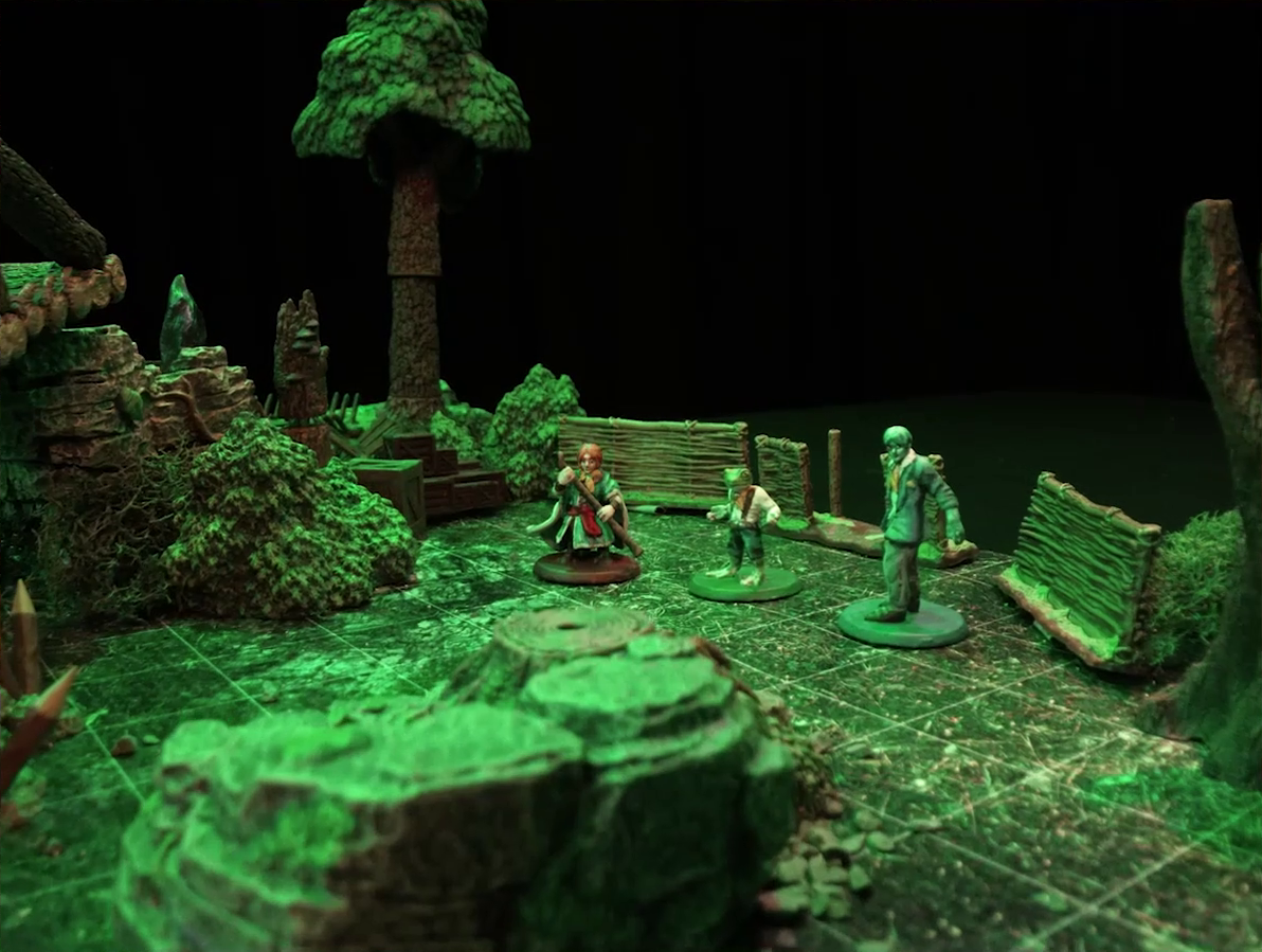 A battlemap of a forest clearing. Sky, Padmund, and Jessep stand in a line behind a rocky outcropping, in front of a fence, and next to some wooden boxes and crates. At the very left of the frame is the edge of a log bridge raised above the forest floor. The bridge begins at a stone ledge.