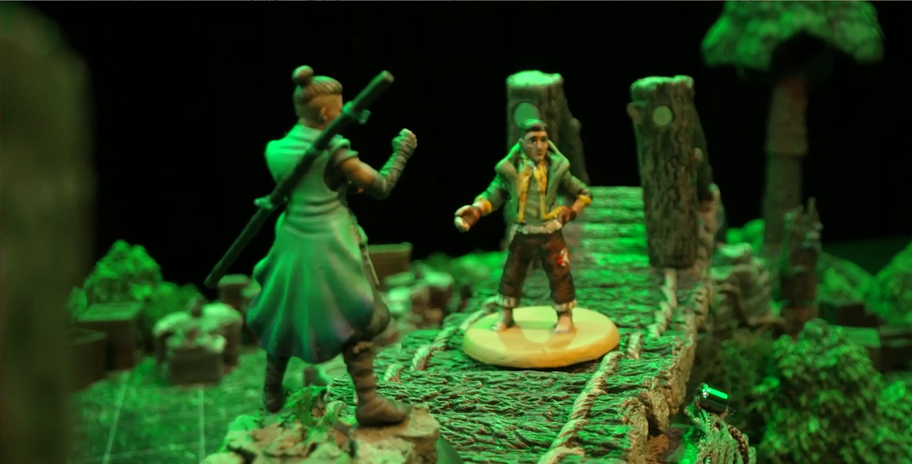 A battlemap focusing on miniatures for Beau and Kai. They stand on a log bridge over a forest floor, on which trees and wooden crates are visible. Beau wears a sleeveless robe with a staff on her back, her hair tied up in a bun. Kai wears ratty brown pants and a green collared jacket with yellow accents.