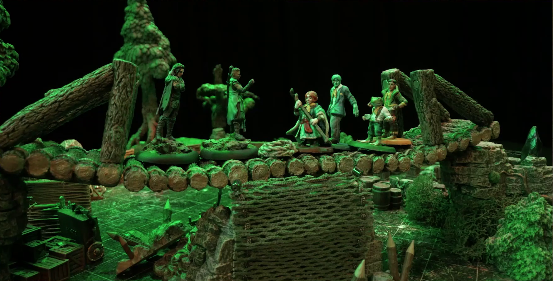 A battlemap focusing on a log bridge spanning two stone ledges in a forest clearing. On the bridge from left to right are Yasha, Beau, Sky, Jessep, Padmund, and Kai. Yasha and Beau face off against the others. A coil of rope is in the center of the bridge. A net of climbable-looking ropes leads up from the ground to the bridge’s center. A number of crates, barrels, and wooden stakes are skattered on the forest floor below the bridge.