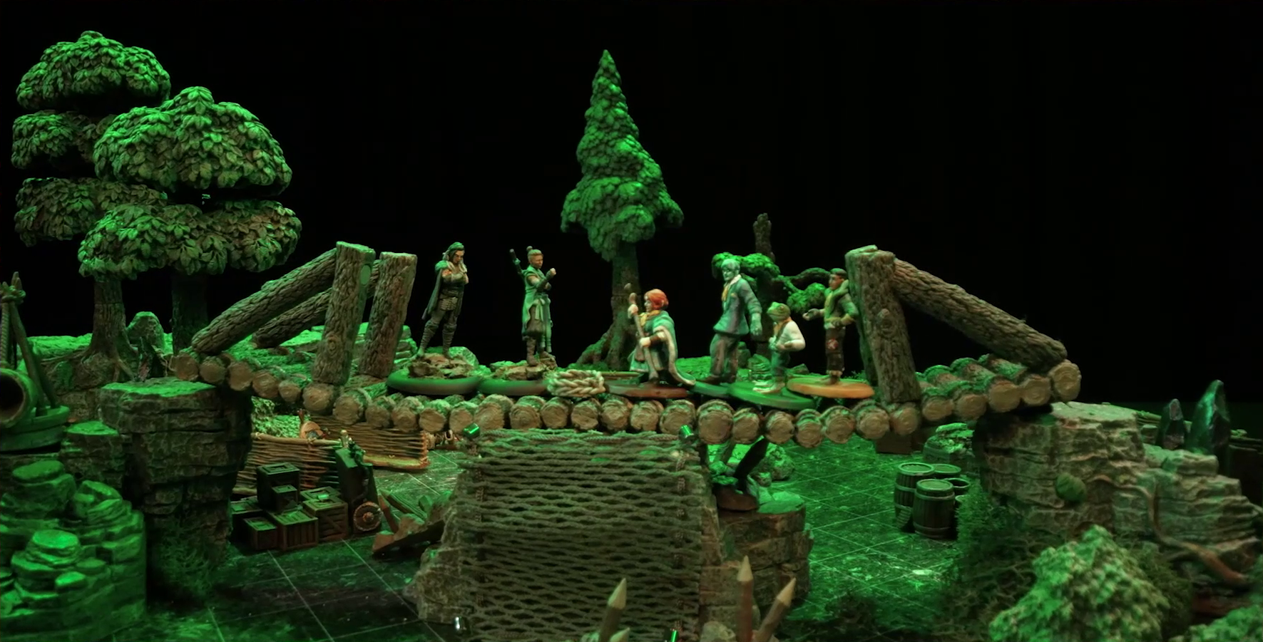 A battlemap focusing on a log bridge spanning two stone ledges in a forest clearing. On the bridge from left to right are Yasha, Beau, Sky, Jessep, Padmund, and Kai. Yasha and Beau face off against the others. A coil of rope is in the center of the bridge. A net of climbable-looking ropes leads up from the ground to the bridge’s center. A number of crates, barrels, and wooden stakes are skattered on the forest floor below the bridge.