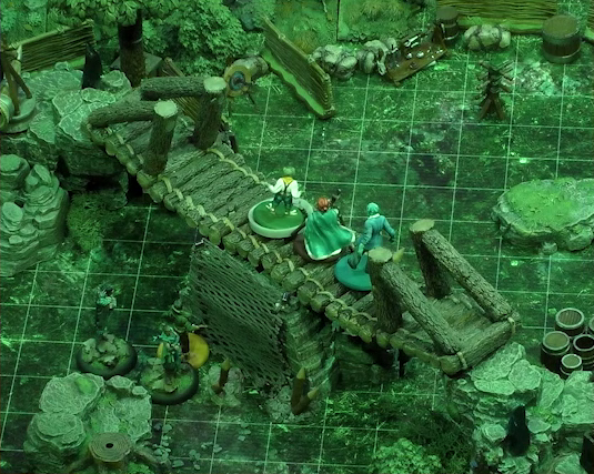 A battlemap of a forest clearing focusing on a log bridge spanning two stone ledges raised above the forest floor. Padmund, Sky, and Jessep stand on the bridge. Yasha, Beau, and Kai are on the ground to the side of the bridge, near a net of climbable-looking ropes leading up to the bridge surface. Some barrels and an archery target are visible on the ground.