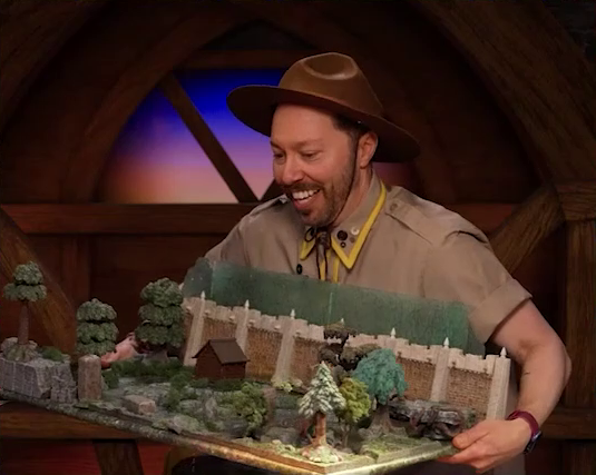 Sam carrying a battlemap of a wooded courtyard bordered on one side by a tall stone defensive wall. A translucent blue magic-looking barrier tops the wall. A small wooden shed sits in the center of the courtyard. Sam wears a tan scout uniform and brown brimmed campaign hat.