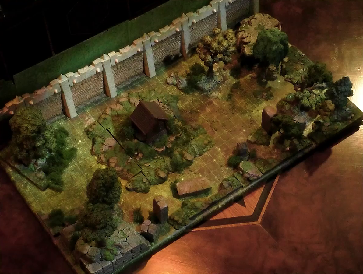 A battlemap of a wooded courtyard bordered on one side by a tall stone defensive wall. A translucent blue magic-looking barrier tops the wall. A small wooden shed sits among some bushes in the center of the courtyard. Filling the rest of the space are trees and old-looking standing stones with circular runic patterns carved into their sides.