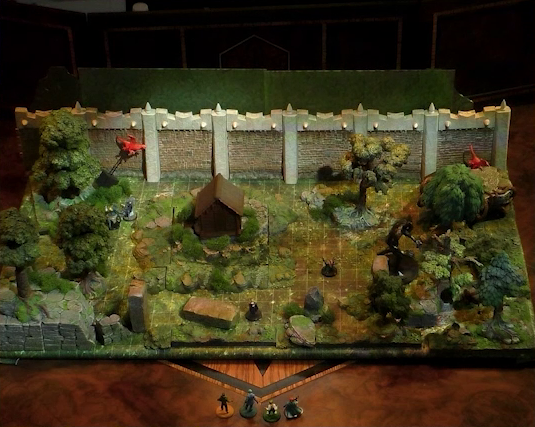 A battlemap of a wooded courtyard bordered on one side by a tall stone defensive wall. A translucent blue magic-looking barrier tops the wall. A small wooden shed sits among some bushes in the center of the courtyard. A number of creatures, including two flying red beasts and a large jet-black humanoid, are spread throughout the courtyard. Kai, Jessep, Padmund, and Sky stand just off the edge of the battlemap looking in. Filling the rest of the space are trees and old-looking standing stones with circular runic patterns carved into their sides.