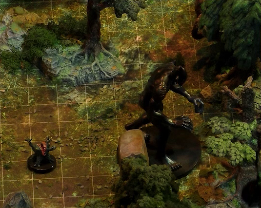 A close-up of two monstrous miniatures in a wooded battlemap. One is a large jet-black humanoid with no clothing and no discernable face, lurching forward with its hands out. The other is a small ball-shaped creature with its torso split open in a toothy maw, two clawed hands emerging from its headless body. The body is held up by four stubby legs.