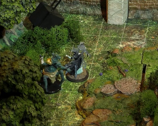 A close-up of two monstrous miniatures in a wooded battlemap. One has teal skin and four spider-like legs propping up a torso with two arms and a rodent-like head. It wears a silver bracer and gold pauldron on one arm and a loincloth around its waist. The other seems to be a robed humanoid surrounded by thick translucent strands.