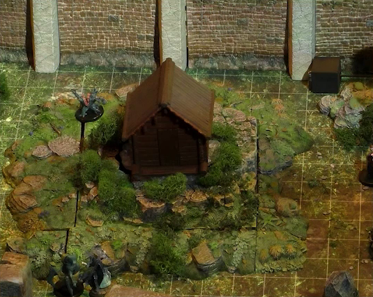 A close-up of a battlemap showing a section of a wooded courtyard with a stone defensive wall at the top edge of the image. A small wooden shed sits among some bushes in the center of the courtyard. Around the shed are three monsters. One is a small ball-shaped creature with its torso split open in a toothy maw, two clawed hands emerging from its headless body. The second has teal skin and four spider-like legs propping up a torso with two arms and a rodent-like head. The third seems to be a robed humanoid surrounded by thick translucent strands.
