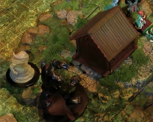A close-up of two monstrous miniatures in front of a small wooden shed on a wooded battlemap. One is a pillar of swirling wind with a subtly-visible open-mouthed face on the front and the rough shape of an arm on the side. The other is a large jet-black humanoid with no clothing and no discernable face, lurching forward with its hands out. Jessep and Sky are just visible behind the shed near a downed ball-shaped creature with its torso split open in a toothy maw, two clawed hands emerging from its headless body.