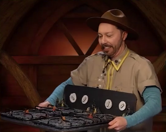 Sam carrying a battlemap that looks like a stovetop. Small flames sprout from each of the four burners and four knobs are on the back panel. Sam wears a tan scout uniform and brown brimmed campaign hat.