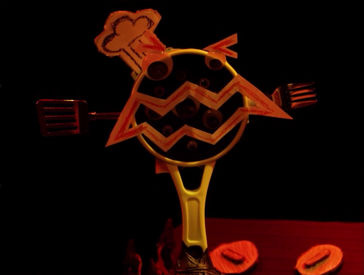 A close-up of a miniature of Wok Wok, the giant frying pan creature. It stands up on its handle. Its mouth, eyebrows, and chef’s hat are made from cut-up paper. It has two googly eyes and several more in the darkness behind its mouth. Its right arm is a spatula and its left arm is a fork. It stands on a battlemap that looks like a stove top, flame and two knobs visible at the bottom of the image.