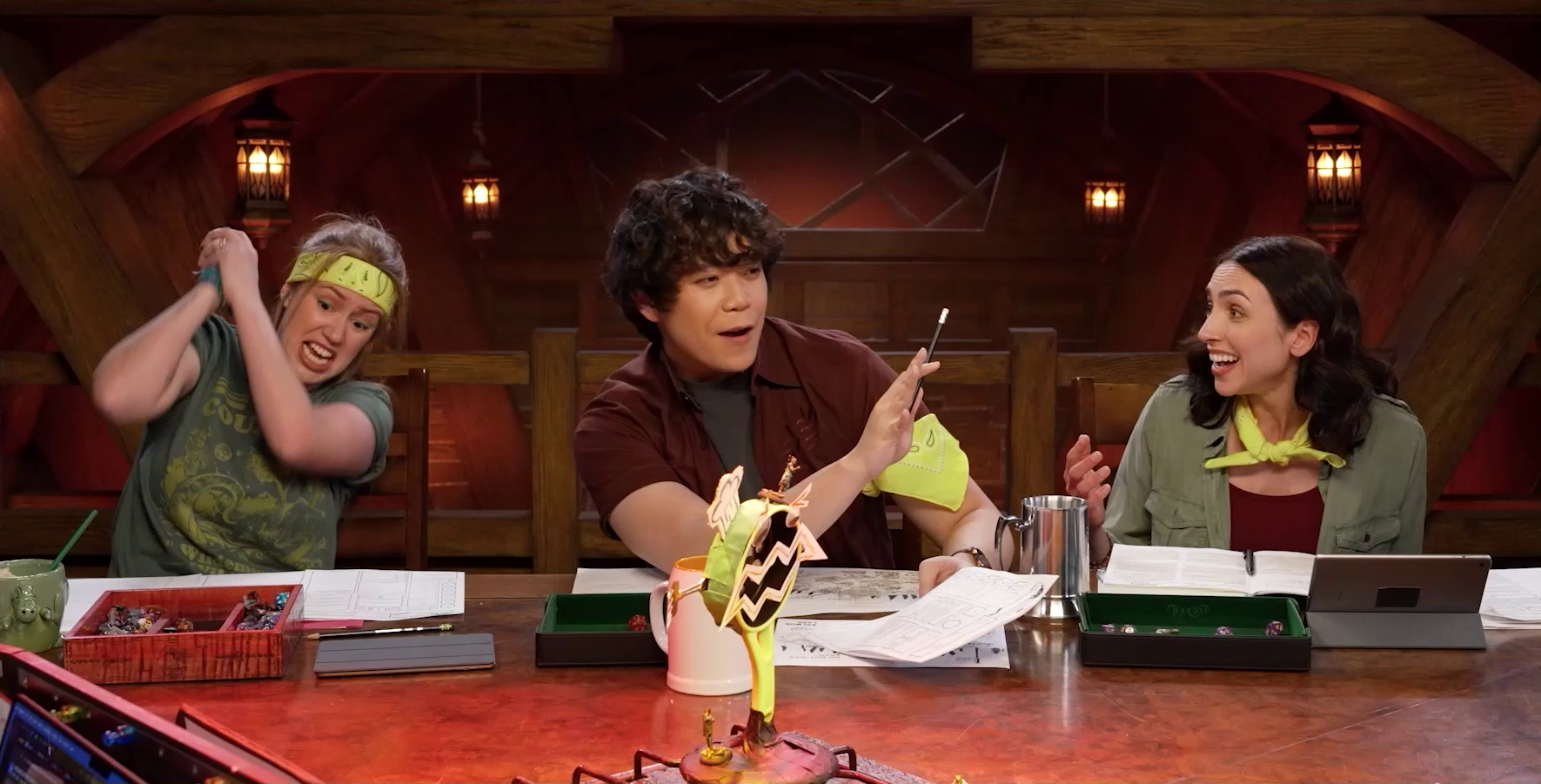 Marisha, Aleks Le, and Eden at the table wearing their yellow Wildemount Wildlings bandanas. Marisha is miming hitting something with a weapon, Aleks is speaking and holding out his hand for emphasis, and Eden is looking at him smiling. In the foreground is visible the edge of a battlemap that looks like a stovetop with Wok Wok, the giant frying pan creature, at its corner burner. Beau is on Wok Wok’s head and Yasha is on its base.