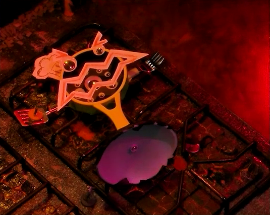 A battlemap that looks like a stovetop. Wok Wok, the giant frying pan creature, lies on its back on the right two burners. Jessep stands between it and the front left burner. Kai stands on Wok Wok’s right spatular hand. Small flames sprout from each burner. The base of the battlemap looks like magma and the scene is bathed in red light.